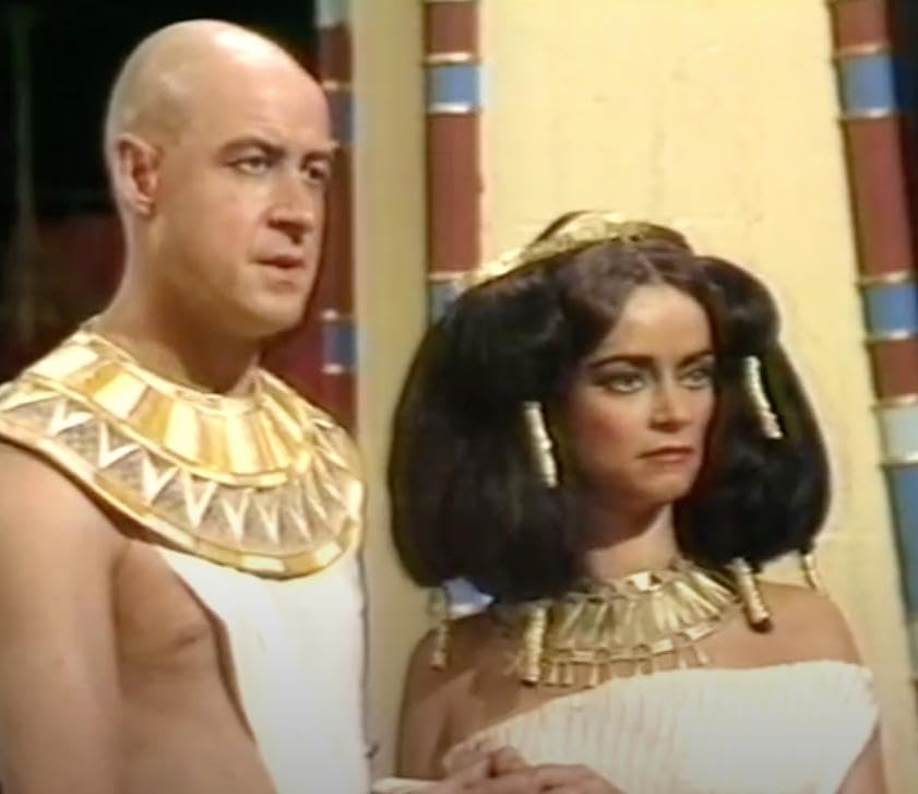 An Egyptian man with a shaved head and a nipple popping out of his costume, and a woman in a massive wig.